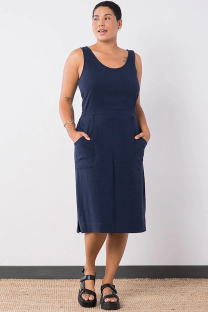 Woman wearing the Jennifer Glasgow Design Aisha Dress in Navy, a sleeveless scoop neck midi dress with a seamed waist and deep patch pockets, with both hands in the pockets and black sport sandals, against a white studio wall.