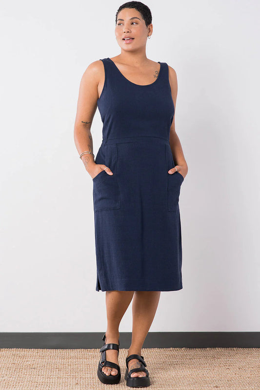 Woman wearing the Jennifer Glasgow Design Aisha Dress in Navy, a sleeveless scoop neck midi dress with a seamed waist and deep patch pockets, with both hands in the pockets and black sport sandals, against a white studio wall.