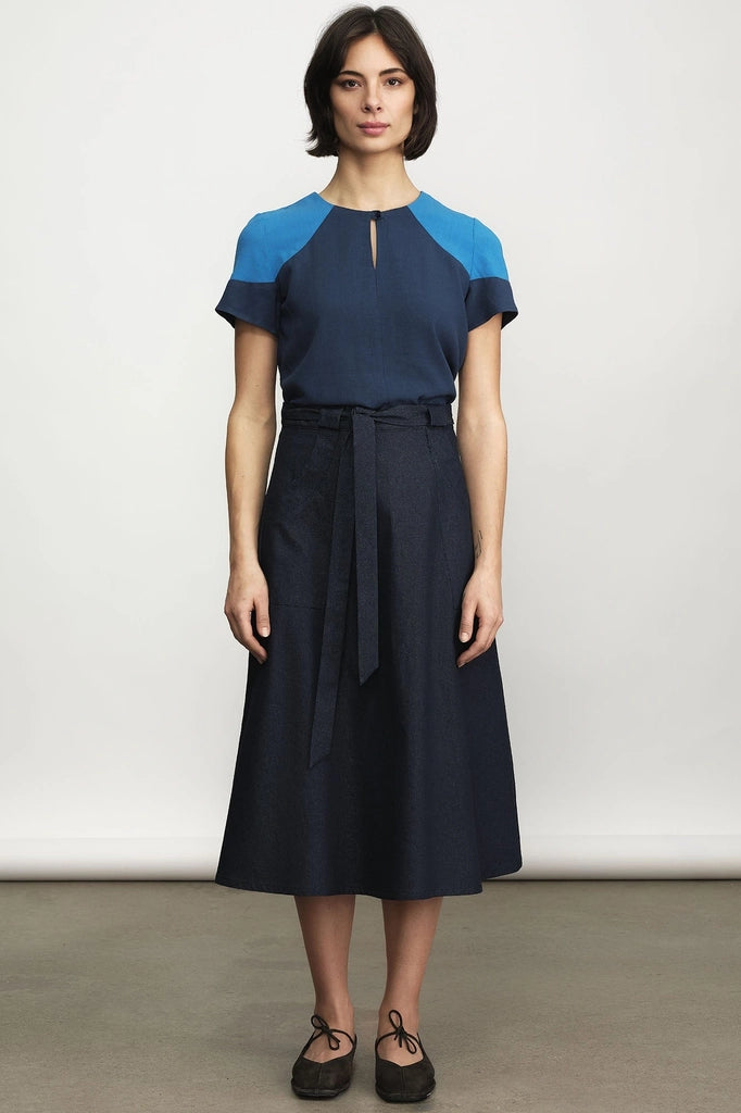 Woman wearing the Jennifer Glasgow Design Saskia Skirt in dark denim with a tie waist, paired with a navy and bright blue colour-block top and black lace-up flats, against a light grey studio background.
