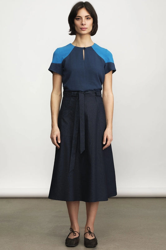 Woman wearing the Jennifer Glasgow Design Saskia Skirt in dark denim with a tie waist, paired with a navy and bright blue colour-block top and black lace-up flats, against a light grey studio background.