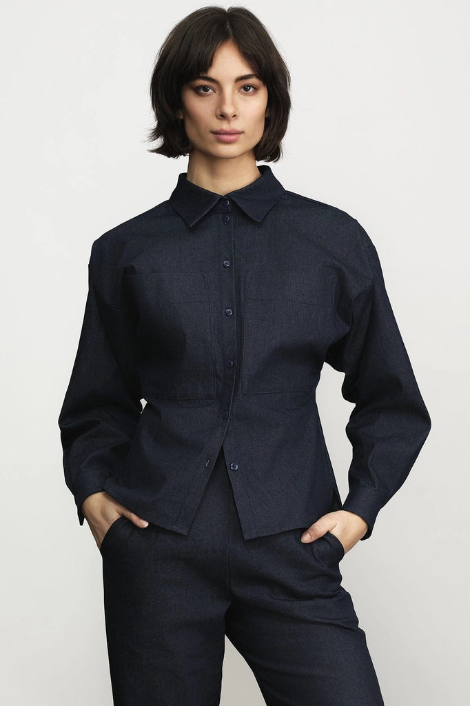 Woman wearing the Jennifer Glasgow Design Tallulah Blouse in dark denim, a long-sleeve button-up with a pointed collar and cinched waist, with matching denim trousers and her hands in her pockets, against a light grey studio background. Available at Textile Apparel in Kelowna.