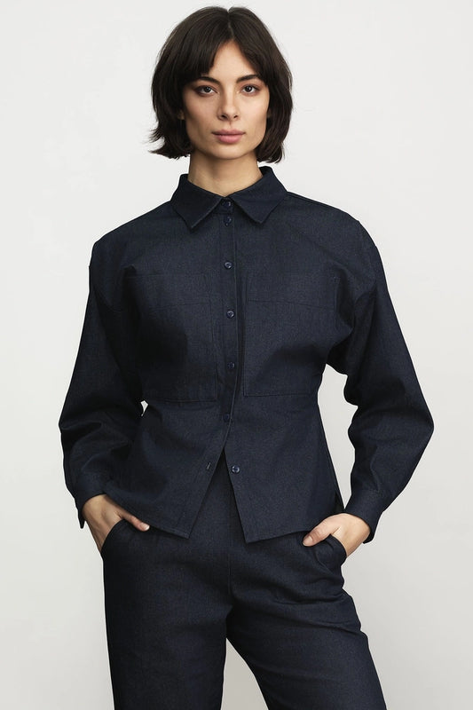 Woman wearing the Jennifer Glasgow Design Tallulah Blouse in dark denim, a long-sleeve button-up with a pointed collar and cinched waist, with matching denim trousers and her hands in her pockets, against a light grey studio background. Available at Textile Apparel in Kelowna.
