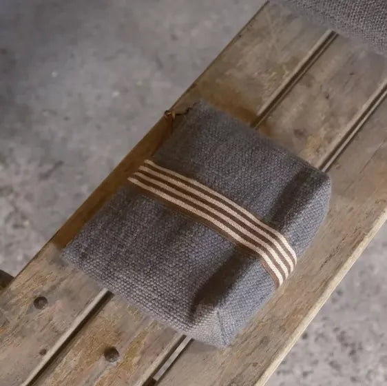 Libeco Jasper Pouch in charcoal grey woven linen and wool with a vertical cream and brown striped band and a long brown zip pull layed on a wooden bench.