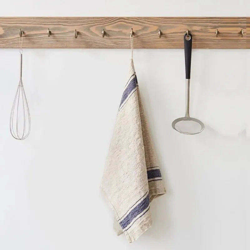 A wooden kitchen hook rack mounted on a white wall, with a wire whisk, a striped beige kitchen towel, and a metal ladle hanging from the hooks.