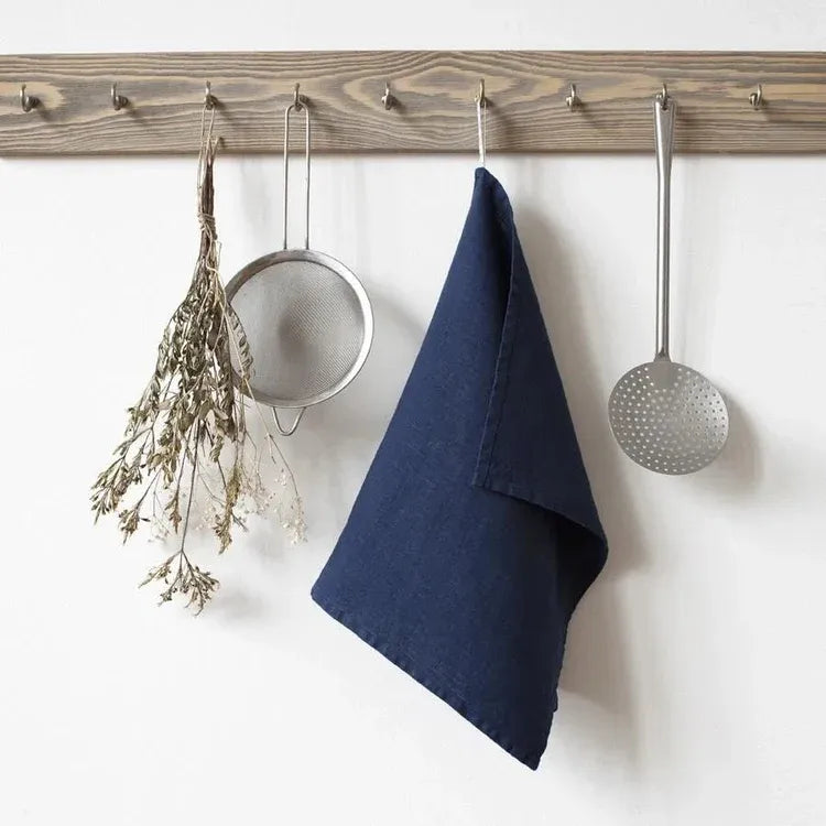 Wooden peg rack with hooks holding a blue towel, metal strainer, and metal ladle on a white wall.