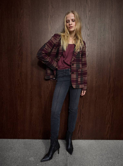 A woman stands against a dark wood-paneled wall wearing a burgundy and brown plaid blazer over a burgundy top, dark gray skinny jeans, and black pointed-toe heeled ankle boots.