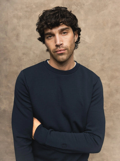 Close-up of a man with folded arms wearing the MOS MOSH Gallery Canada Abel Crew Neck Sweat in salute navy melange, against a warm beige textured backdrop.