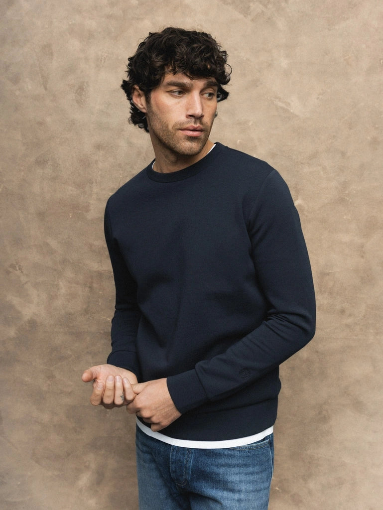 Man wearing the MOS MOSH Gallery Canada Abel Crew Neck Sweat in salute navy melange over a white tee with blue jeans, against a warm beige textured backdrop.