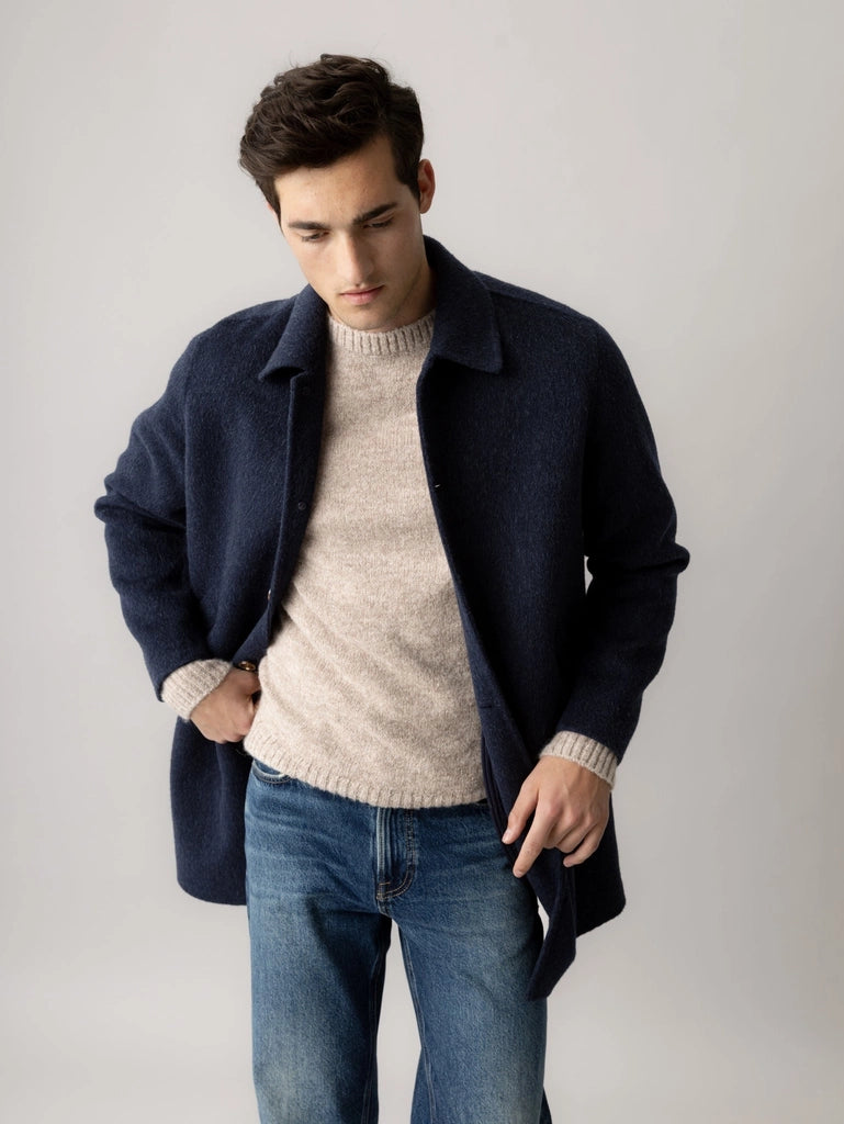 Man looking down and adjusting the MOS MOSH Gallery Canada Barkley Wool Coat in salute navy, worn over an oatmeal knit and jeans, against a light grey studio background.