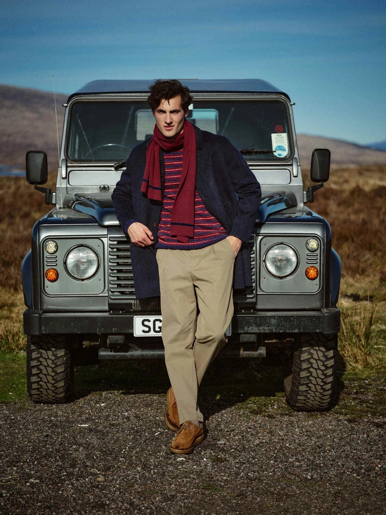 Man leaning against a vintage off-road vehicle wearing the MOS MOSH Gallery Canada Barkley Wool Coat in salute navy, with a burgundy scarf and khaki trousers, in an open moorland landscape.