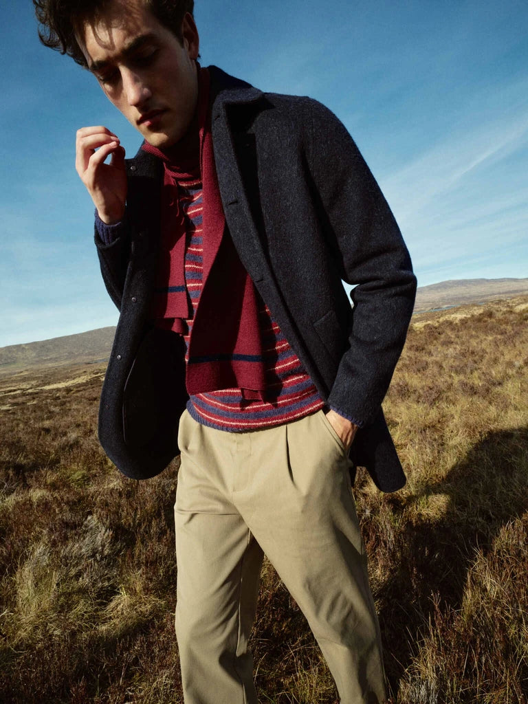 Man standing in tall autumn grass wearing the MOS MOSH Gallery Canada Barkley Wool Coat in salute navy, with a striped knit, burgundy scarf and khaki trousers, under a blue sky.