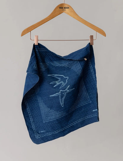 MOS MOSH Gallery Canada Bird Chambray Neckerchief in indigo blue, hanging from a wooden hanger and showing the swallow print and geometric border, against a light grey background.