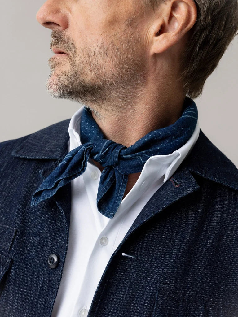 Close-up of the MOS MOSH Gallery Canada Bird Chambray Neckerchief in indigo blue, knotted at a man's neck over a white shirt and denim jacket.