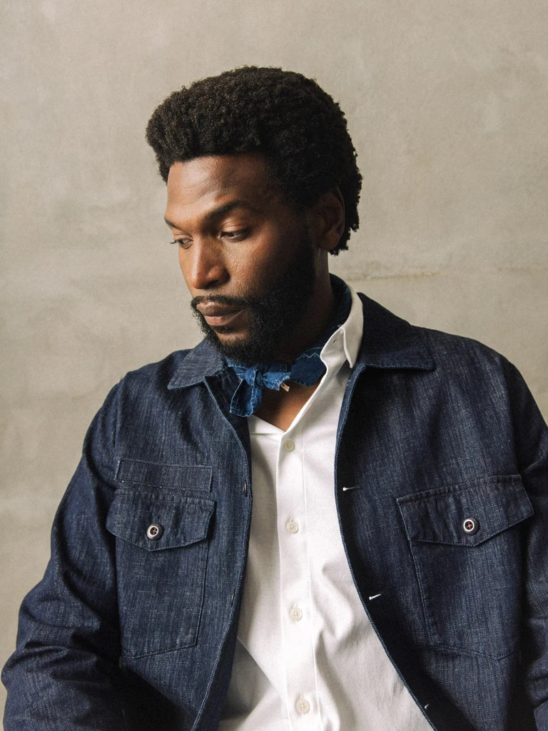 Man wearing the MOS MOSH Gallery Canada Bird Chambray Neckerchief in indigo blue, tied under the collar of a white shirt and a denim jacket, against a warm grey textured wall.
