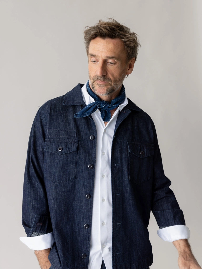 Man wearing the MOS MOSH Gallery Canada Bird Chambray Neckerchief in indigo blue, knotted at the neck with a white shirt and dark denim overshirt, against a light grey studio background.