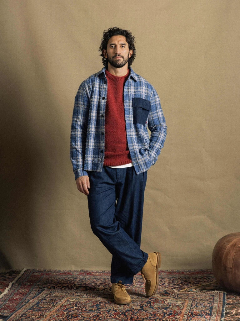 Man standing on a patterned rug wearing the MOS MOSH Gallery Canada Carlos Crew Neck Knit in burnt russet under a blue plaid overshirt, with dark denim trousers and tan suede shoes, against a warm beige backdrop.