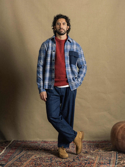 Man standing on a patterned rug wearing the MOS MOSH Gallery Canada Carlos Crew Neck Knit in burnt russet under a blue plaid overshirt, with dark denim trousers and tan suede shoes, against a warm beige backdrop.