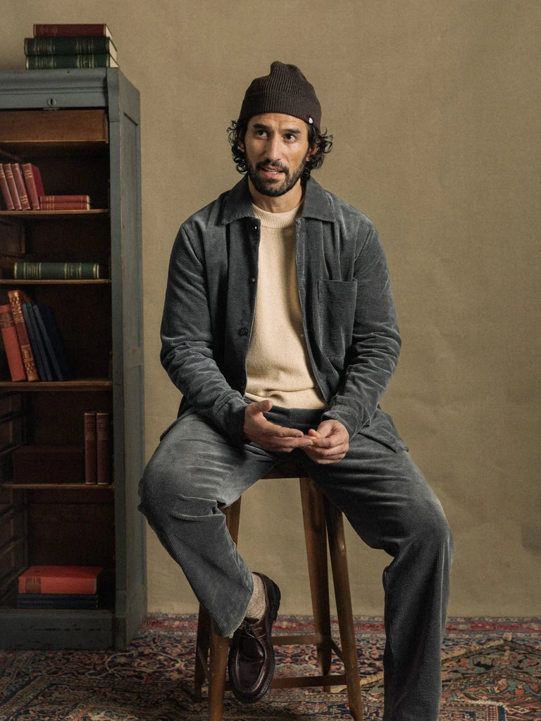 Man wearing MOS MOSH Gallery Canada Conor Corduroy Pant in Castlerock with a matching overshirt, cream knit and knit hat, seated on a wooden stool beside a bookshelf.