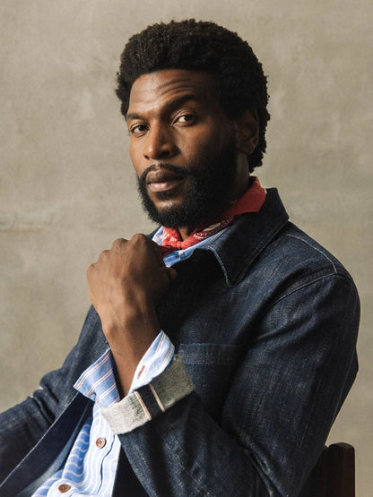 Close-up of the MOS MOSH Gallery Canada Micca Selvedge Jacket in dark blue denim, showing the cuffed sleeve and selvedge edge, worn with a red neckerchief against a textured wall.