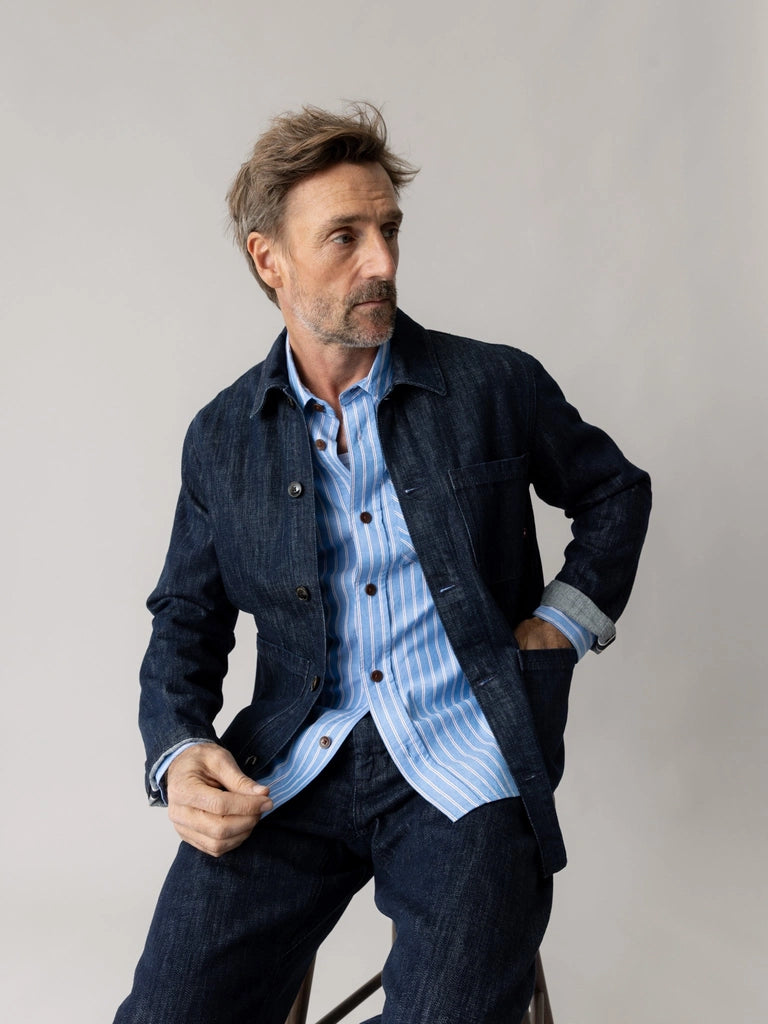 Man sitting on a stool in the MOS MOSH Gallery Canada Micca Selvedge Jacket in dark blue denim, with a striped blue shirt and the sleeves turned back, against a light grey background.