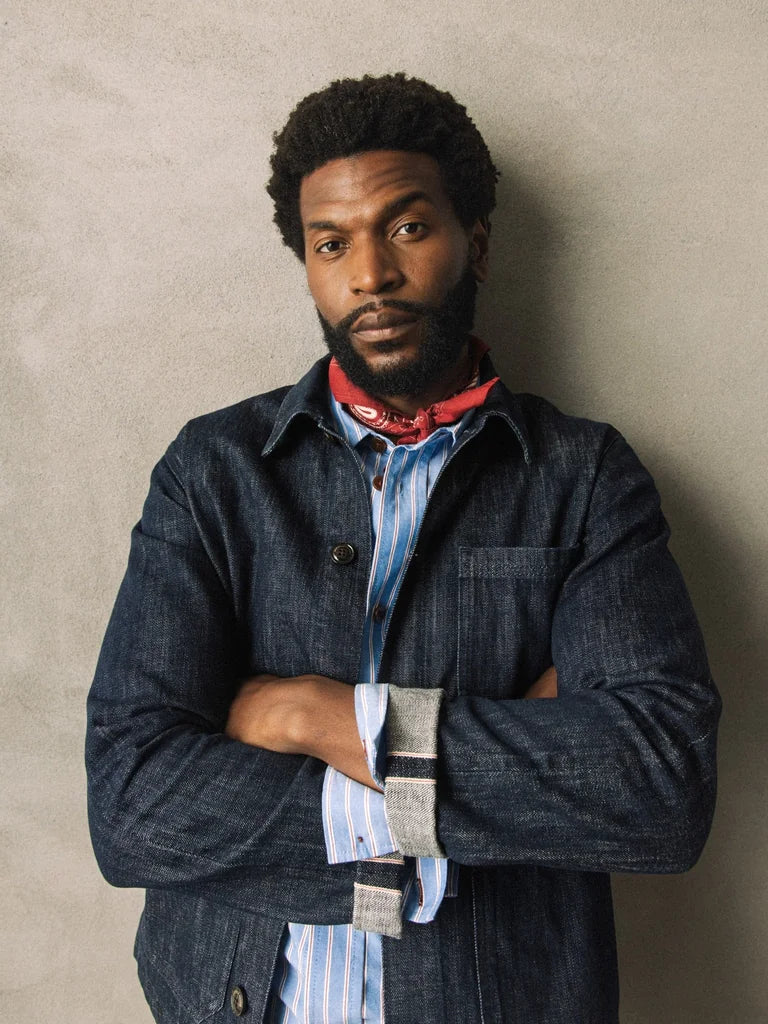 Man with crossed arms wearing the MOS MOSH Gallery Canada Micca Selvedge Jacket in dark blue denim with a striped shirt and a red neckerchief, against a textured stone-coloured wall.