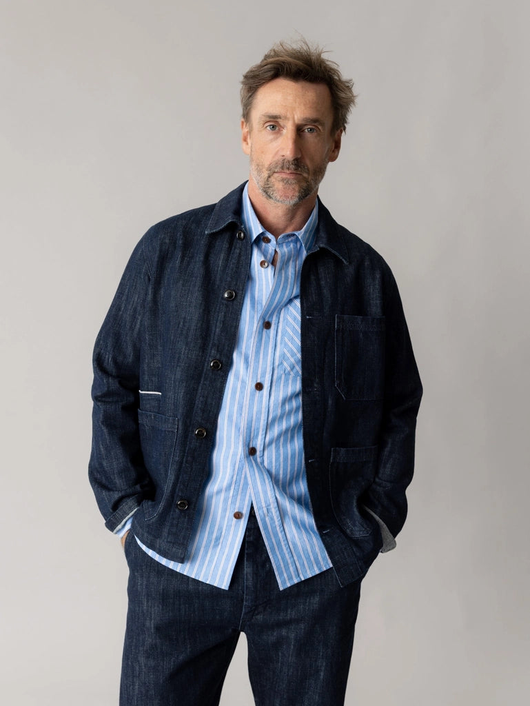 Man wearing the MOS MOSH Gallery Canada Micca Selvedge Jacket in dark blue denim, open over a striped blue shirt with matching denim pants, standing against a light grey studio background.