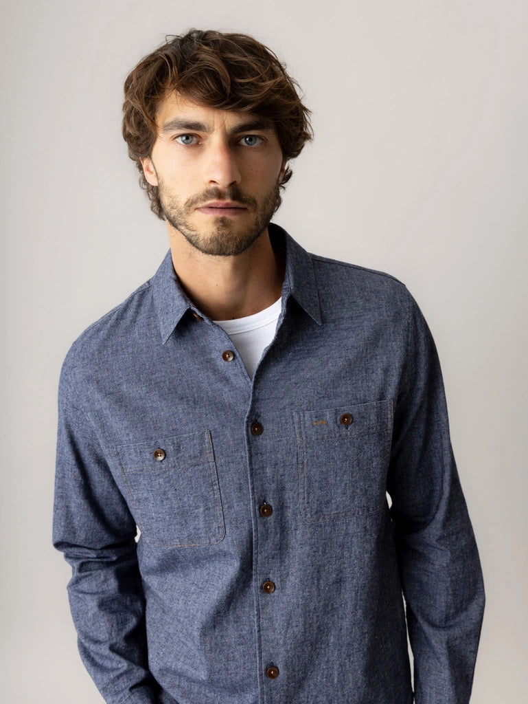 Close-up of a man wearing the MOS MOSH Gallery Canada Norbert Denim Shirt in vintage indigo, showing the chest pockets and brown buttons, against a light grey studio background.
