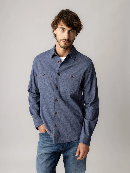 Man wearing the MOS MOSH Gallery Canada Norbert Denim Shirt in vintage indigo, unbuttoned at the collar over a white tee with blue jeans, against a light grey studio background.