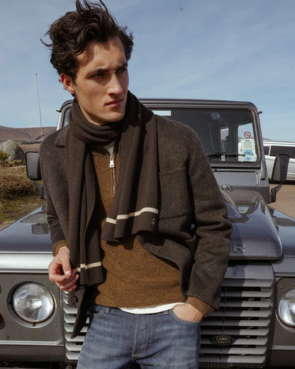 Man leaning against a vintage off-road vehicle wearing the MOS MOSH Gallery Canada Warren Jacket in delicioso brown, with a brown knit, striped scarf and blue jeans, under a blue sky.