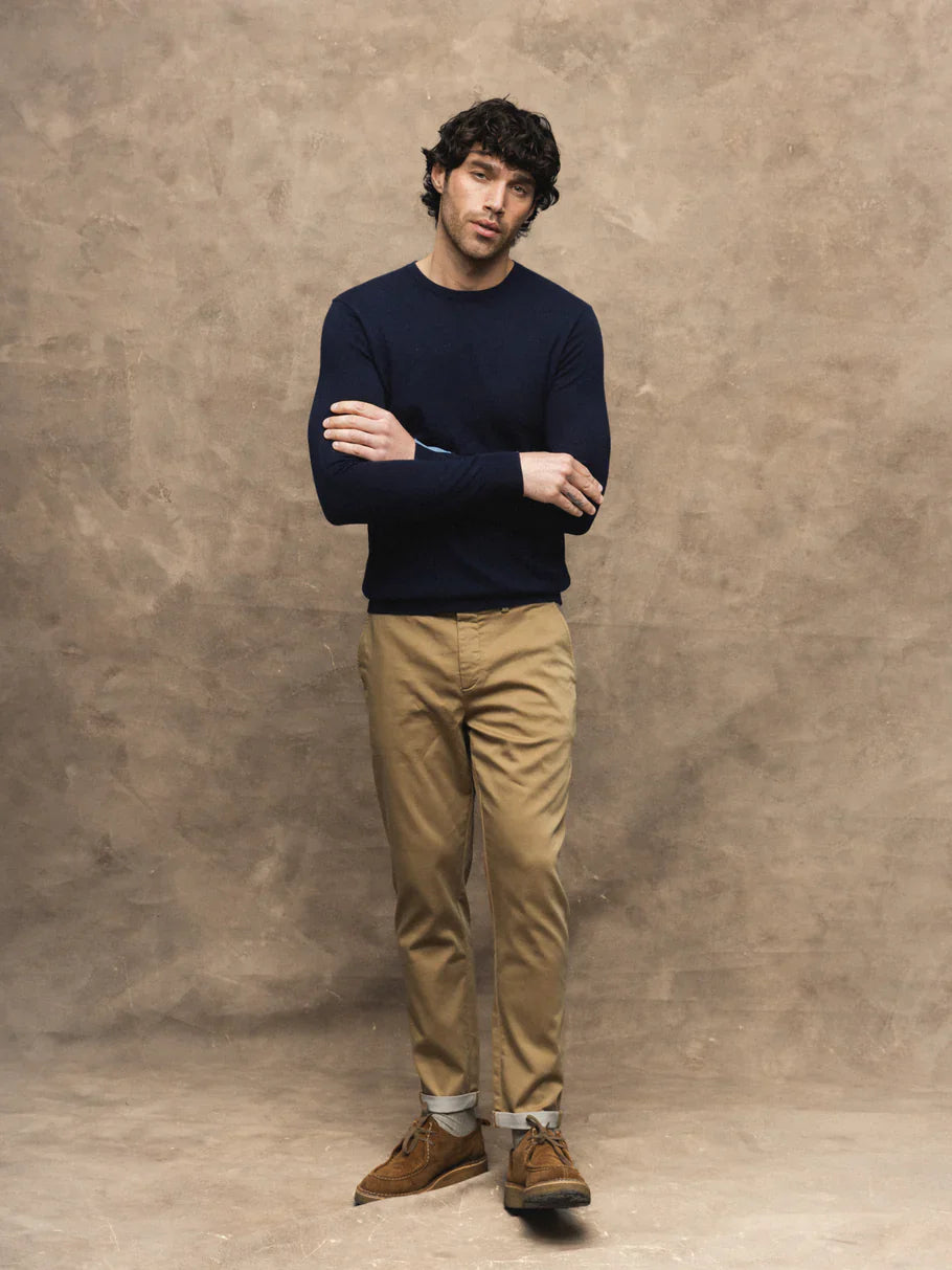 A man stands with arms crossed wearing a dark navy long-sleeve crewneck sweater, tan chinos, and brown suede boots against a textured brown studio backdrop.