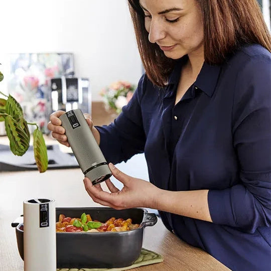 A woman in a navy blue shirt holds a metallic gray electric pepper mill over a dark baking dish with food, with a white version of the mill visible in the foreground on a kitchen counter.