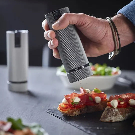 A hand holding a gray cylindrical pepper grinder sprinkling seasoning onto bread topped with prosciutto and mozzarella, with a second pepper grinder visible in the background on a dark tabletop.
