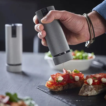 A hand holding a gray cylindrical pepper grinder sprinkling seasoning onto bread topped with prosciutto and mozzarella, with a second pepper grinder visible in the background on a dark tabletop.
