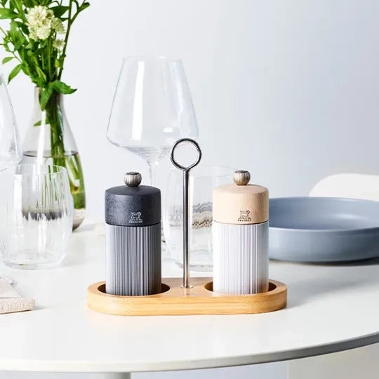 A pair of ribbed salt and pepper mills with one dark gray body and one light beige body, set on a wooden stand with a metal wine glass pick, on a white table with glassware and flowers in the background.