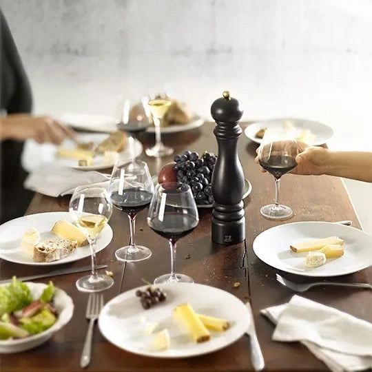A dining table set with white plates, wine glasses containing red and white wine, cheese, grapes, a black pepper mill, and a black salt mill, with partial hands of people visible on both sides.