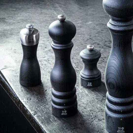 Four dark wooden pepper mills of varying sizes arranged on a textured gray stone surface, with one mill showing a polished silver metal top.