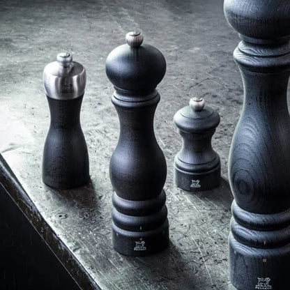 Four dark wooden pepper mills of varying sizes arranged on a textured gray stone surface, with one mill showing a polished silver metal top.