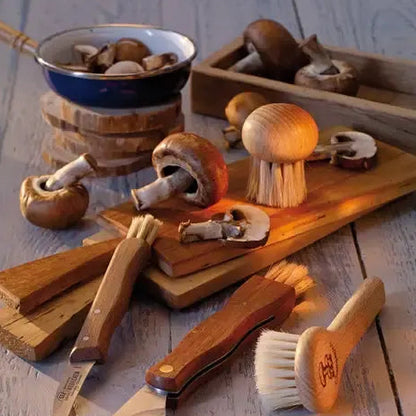 A set of wooden kitchen utensils and tools arranged on a rustic gray wood plank surface, including a small knife, brush, scrubber, cutting boards, and a bowl with mushrooms.