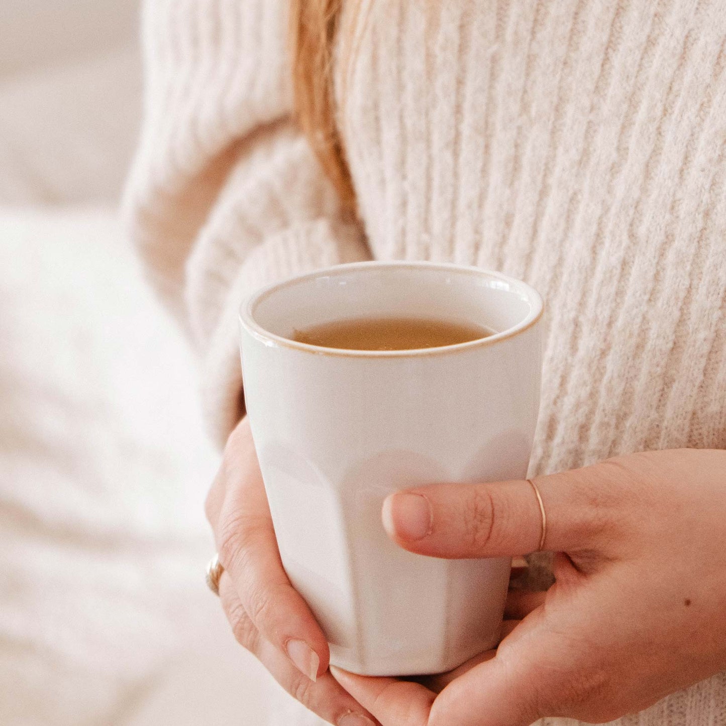 Person holding a white mug with a warm beverage, wearing a beige sweater.