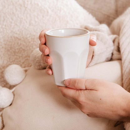 Person holding a white mug with a cozy blanket and pillow in the background