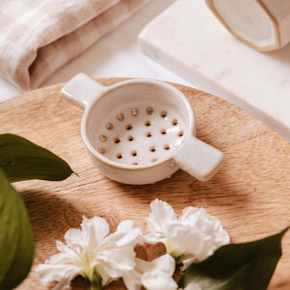 White ceramic strainer on a wooden surface with white flowers and green leaves.