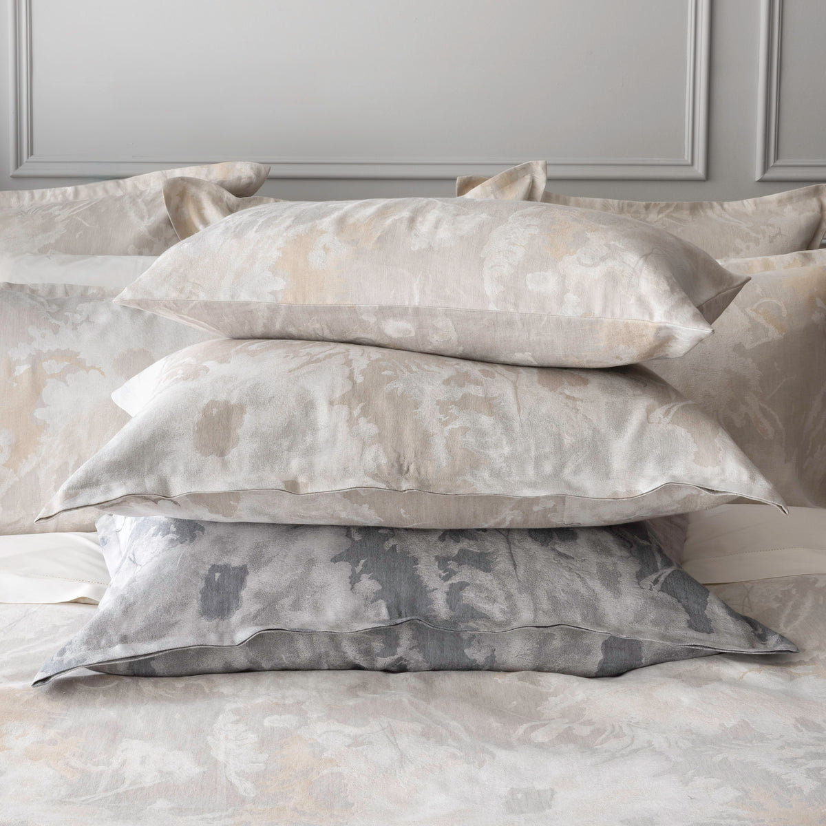 A stack of three Blossom jacquard cushions in Parchment, Oyster and Moonlight on a made bed, showing the knife-edge finish and painterly floral pattern.