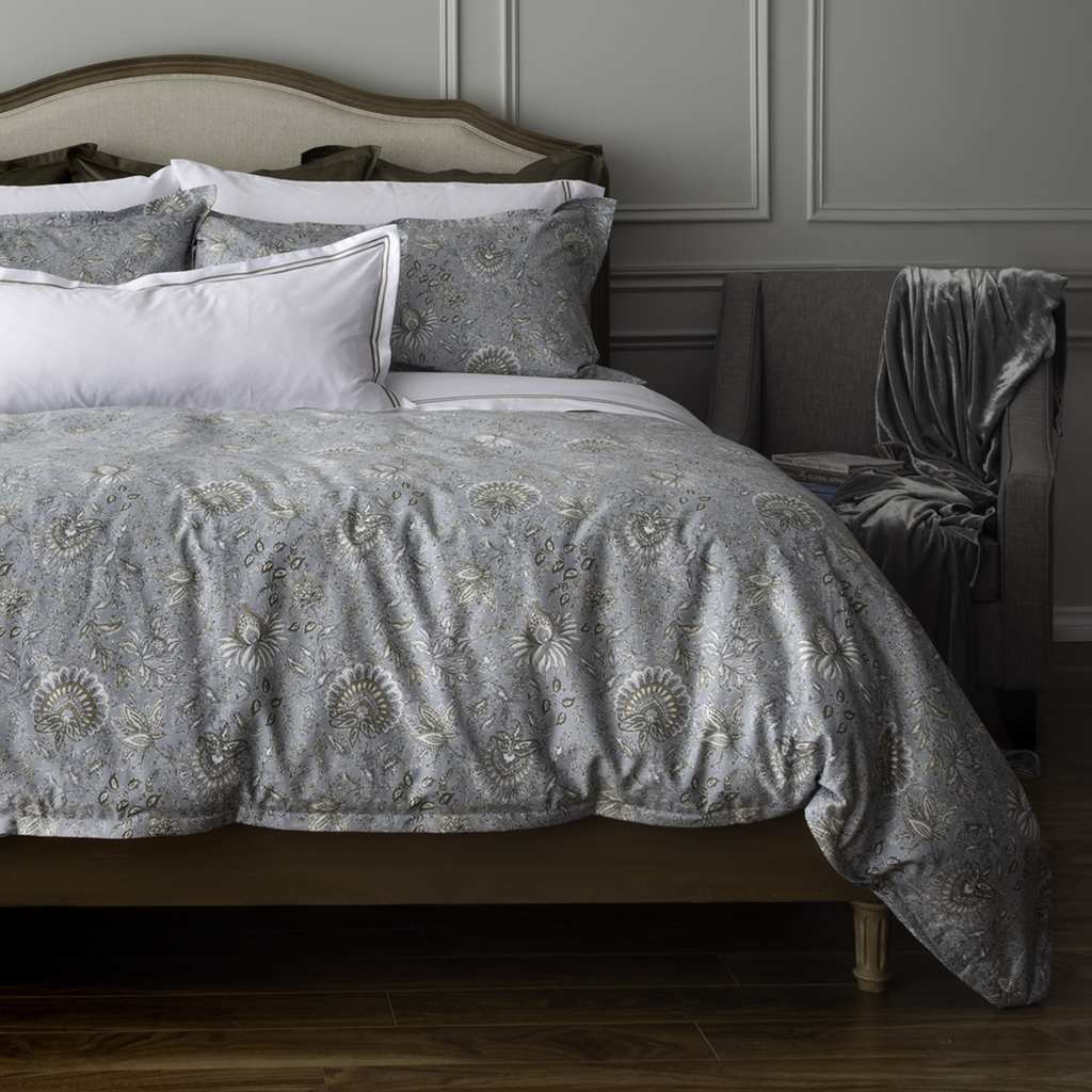 A bed dressed in a gray and beige patterned quilt and matching shams, styled in a bedroom with a wooden headboard, white accent pillows, a gray upholstered chair with a draped throw blanket, and dark wood flooring.