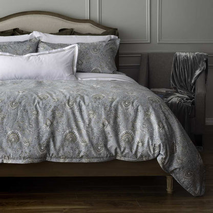 A bed dressed in a gray and beige patterned quilt and matching shams, styled in a bedroom with a wooden headboard, white accent pillows, a gray upholstered chair with a draped throw blanket, and dark wood flooring.