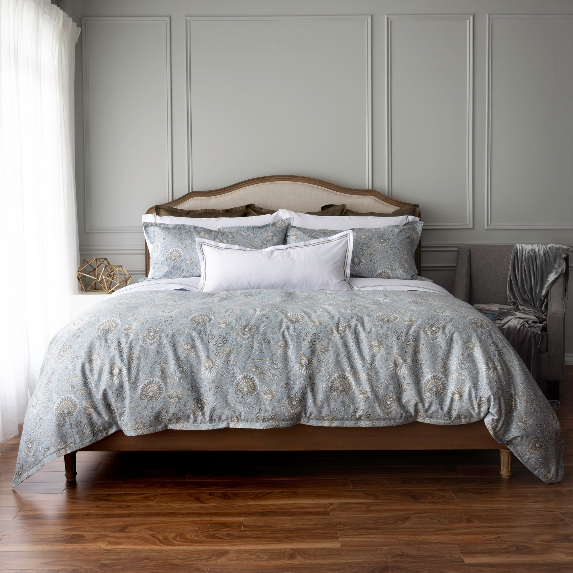 A neatly made bed in a styled bedroom, featuring a light gray and white paisley patterned duvet cover and matching pillows, with a white accent pillow in the center. The room has gray paneled walls, a wooden floor, and a gray armchair with a throw blanket.