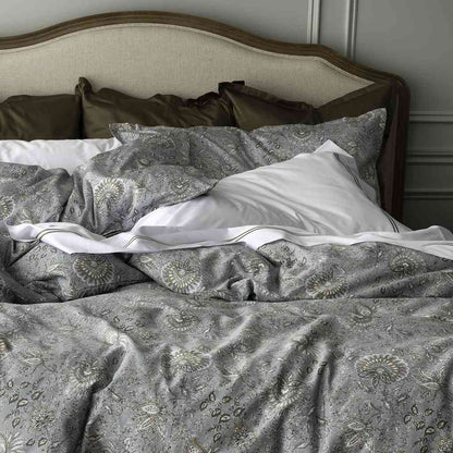 A close-up of a made bed with a patterned duvet cover and matching pillow shams in muted blue-gray and beige, layered with white pillows and olive-brown pillows, set against a wooden upholstered headboard.