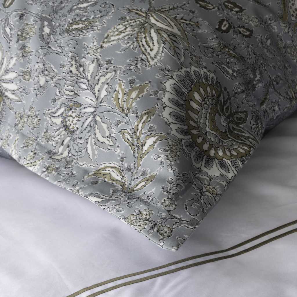 A close-up of a pillow sham with an ornate floral and paisley pattern in muted blue, white, taupe, and gold, resting on white bedding with thin gold stripe accents.