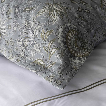 A close-up of a pillow sham with an ornate floral and paisley pattern in muted blue, white, taupe, and gold, resting on white bedding with thin gold stripe accents.