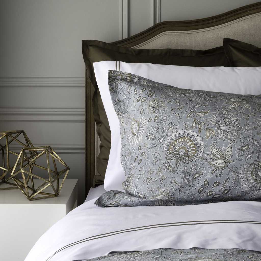 A close-up of a bed styled with layered bedding, featuring a prominent gray damask-patterned pillow in front of white and dark olive pillows, set against a dark wood headboard.