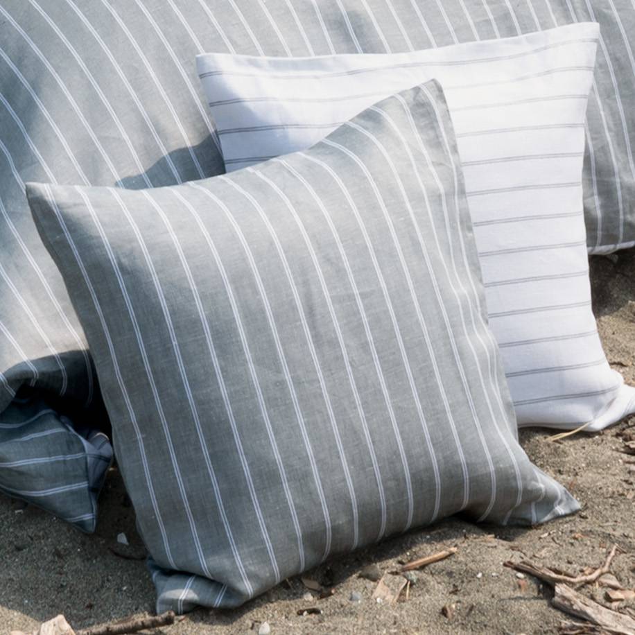 Two striped throw pillows in a linen-like fabric, one in gray and white and one in white and gray, resting on sandy ground outdoors.